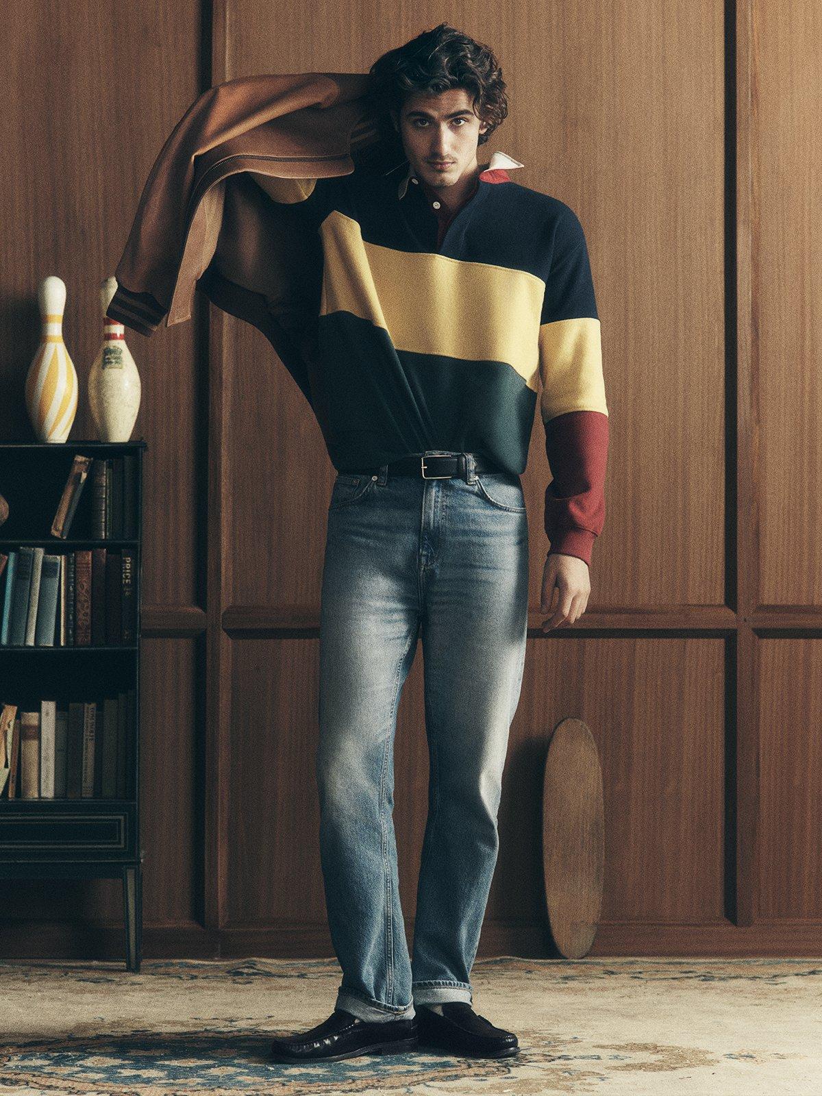 Male model wearing striped rugby shirt, blue jeans, loafers and holding a brown leather varsity Jacket on his shoulder