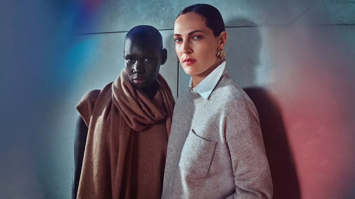 Two people standing staring at the camera one with a scarf on, the other with a white shirt collar coming out of a grey jumper