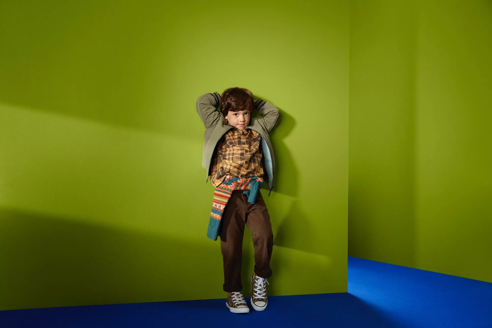 Stylish young boy in casual jacket and trainers, leaning against lime green wall in modern studio setting