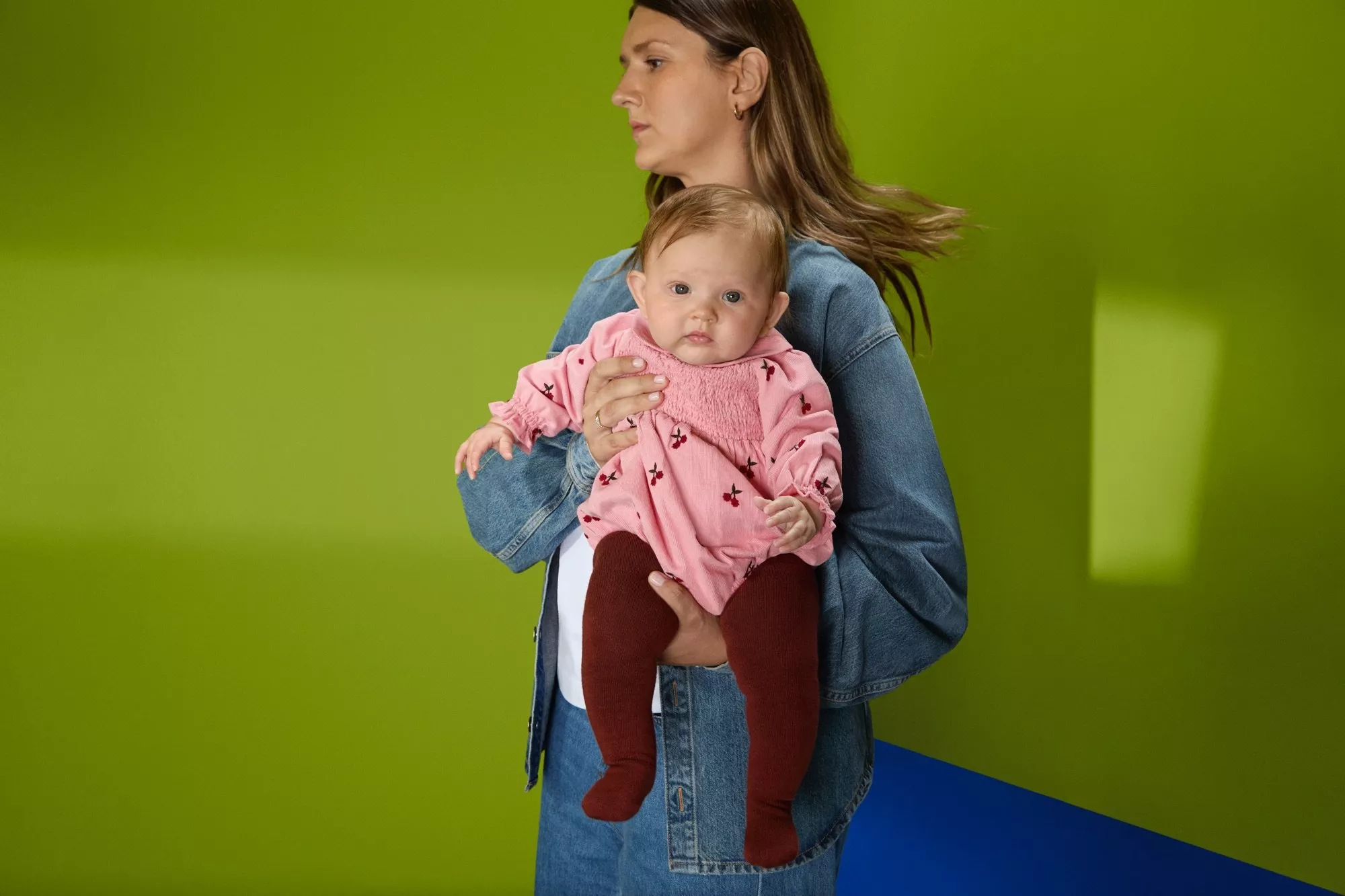 Woman holding baby in pink dress and burgundy tights, studio portrait against bold green background