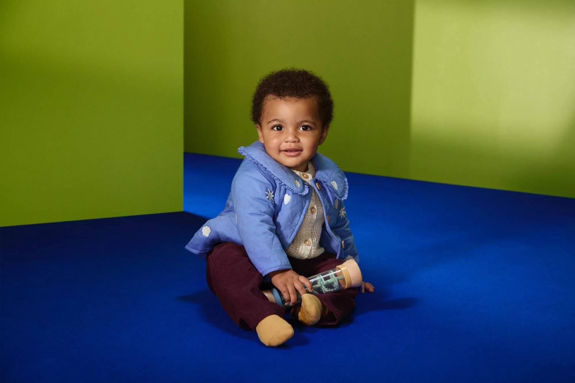 Smiling toddler in blue jacket sitting on a blue sofa, holding a wooden rattle toy against a green wall