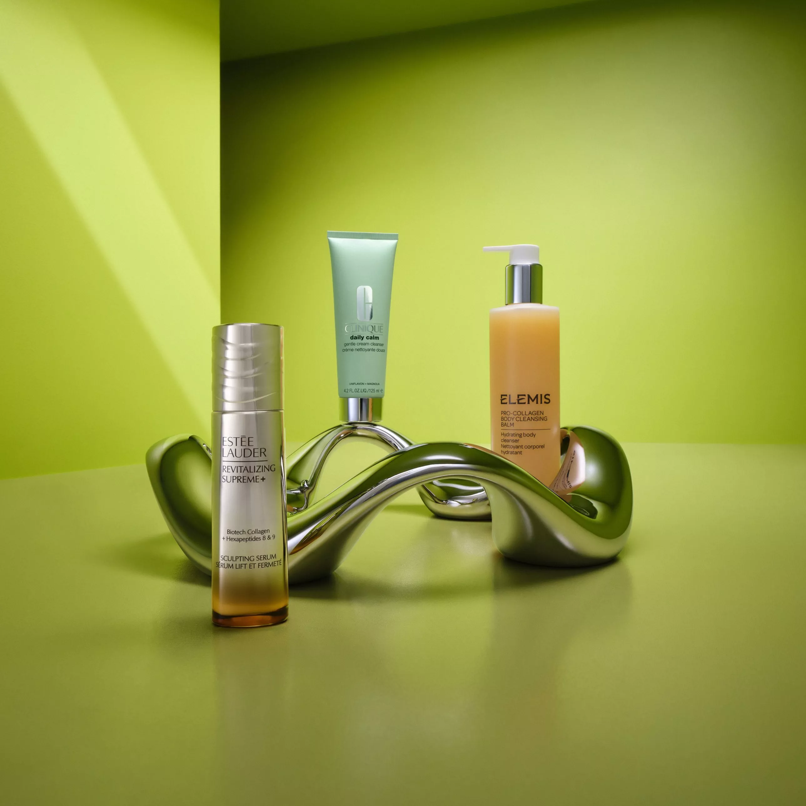 Luxury skincare products on green backdrop, featuring Estée Lauder serum, Clinique cream and Elemis cleanser bottle