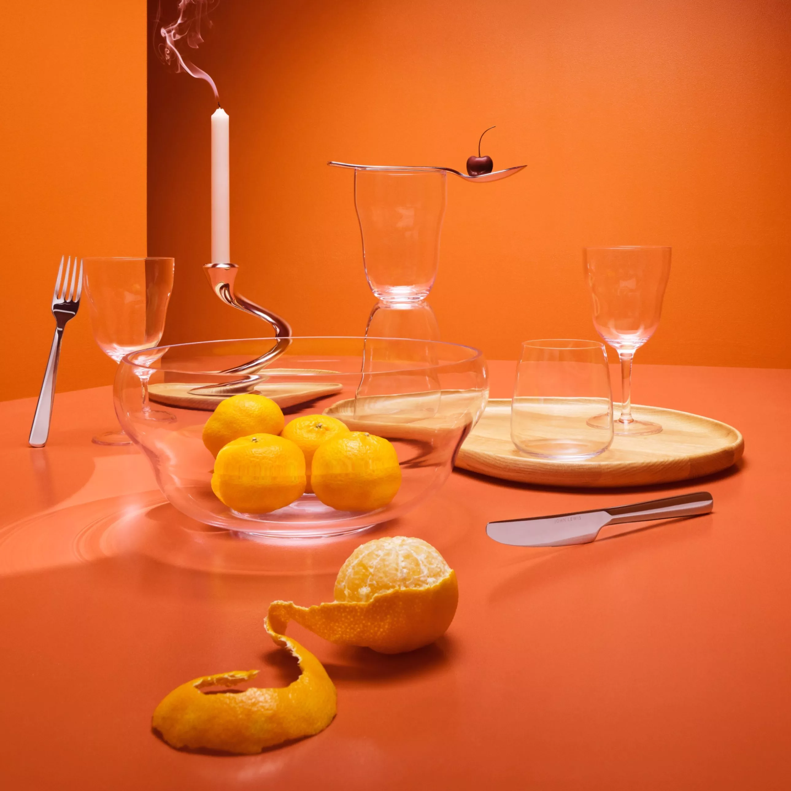 Modern orange still life with glassware, candle, cutlery and citrus fruit on a table setting backdrop
