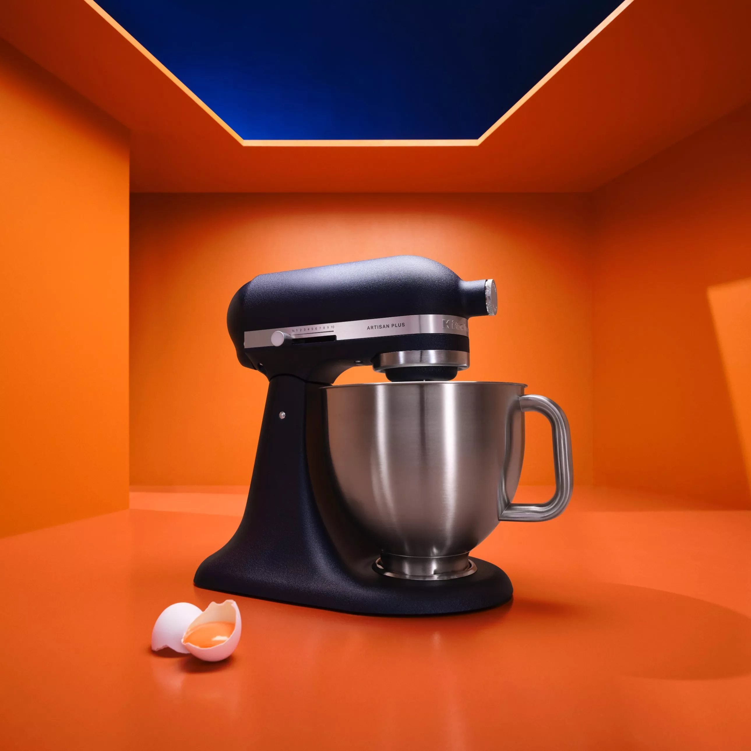 Black stand mixer with stainless steel bowl in orange studio kitchen, with cracked egg shell in foreground