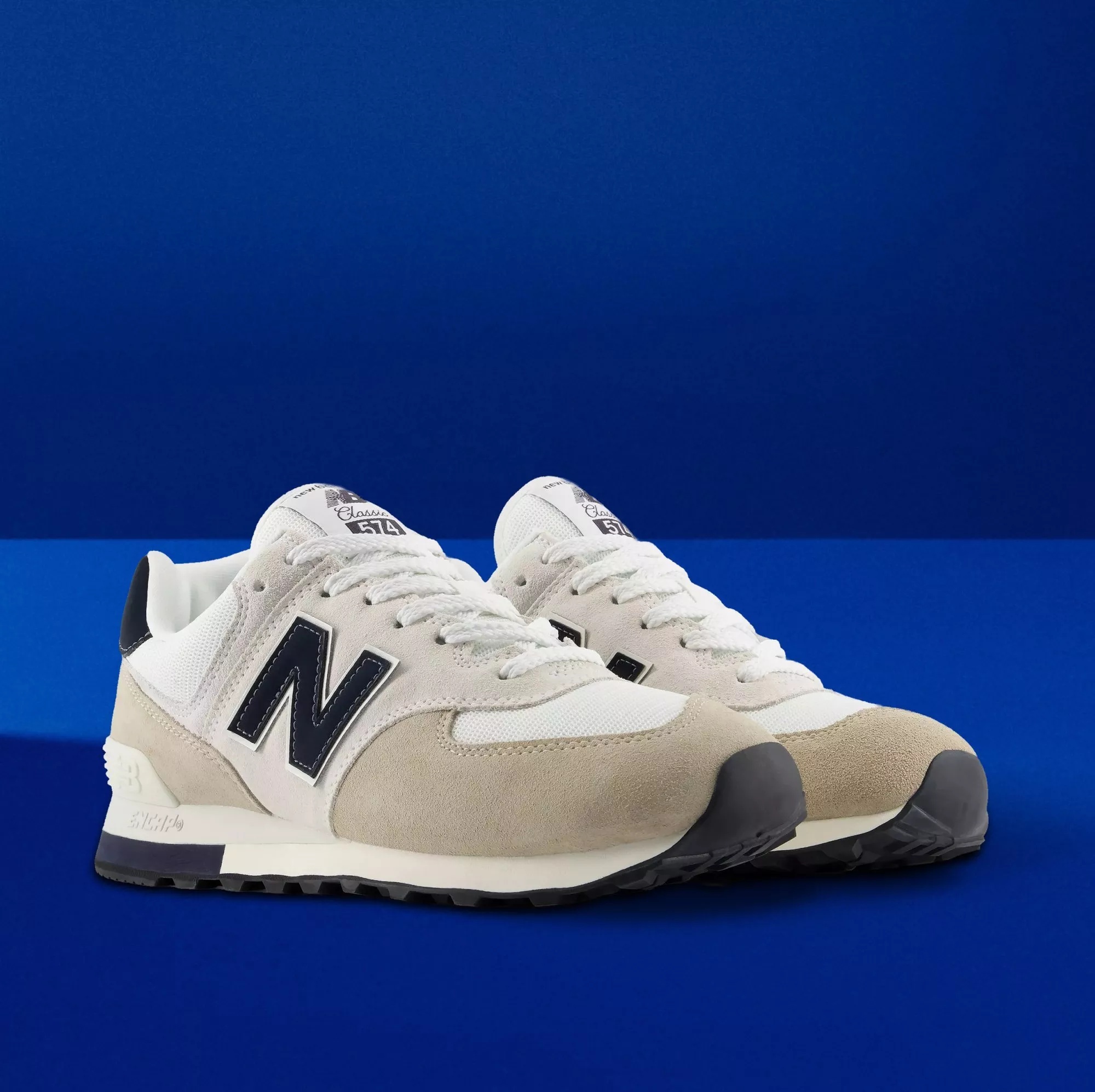 Pair of New Balance 574 trainers in white and beige suede with navy logo, photographed on blue background
