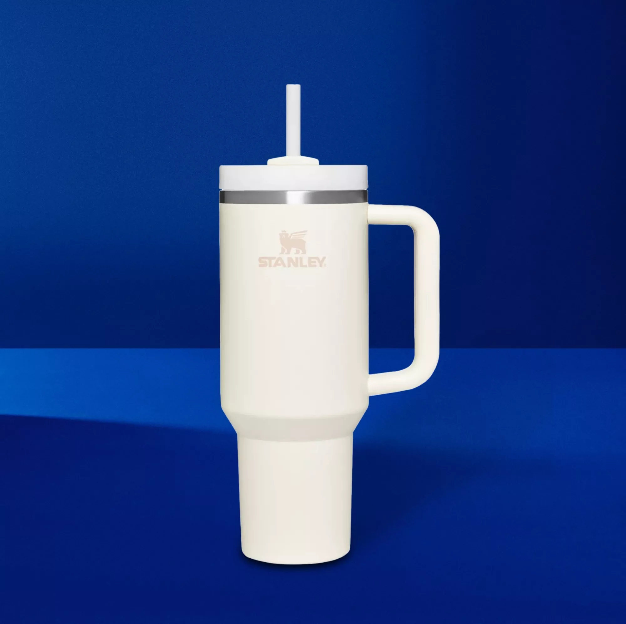 Cream Stanley insulated travel tumbler with handle and straw lid on blue background, reusable drinks cup