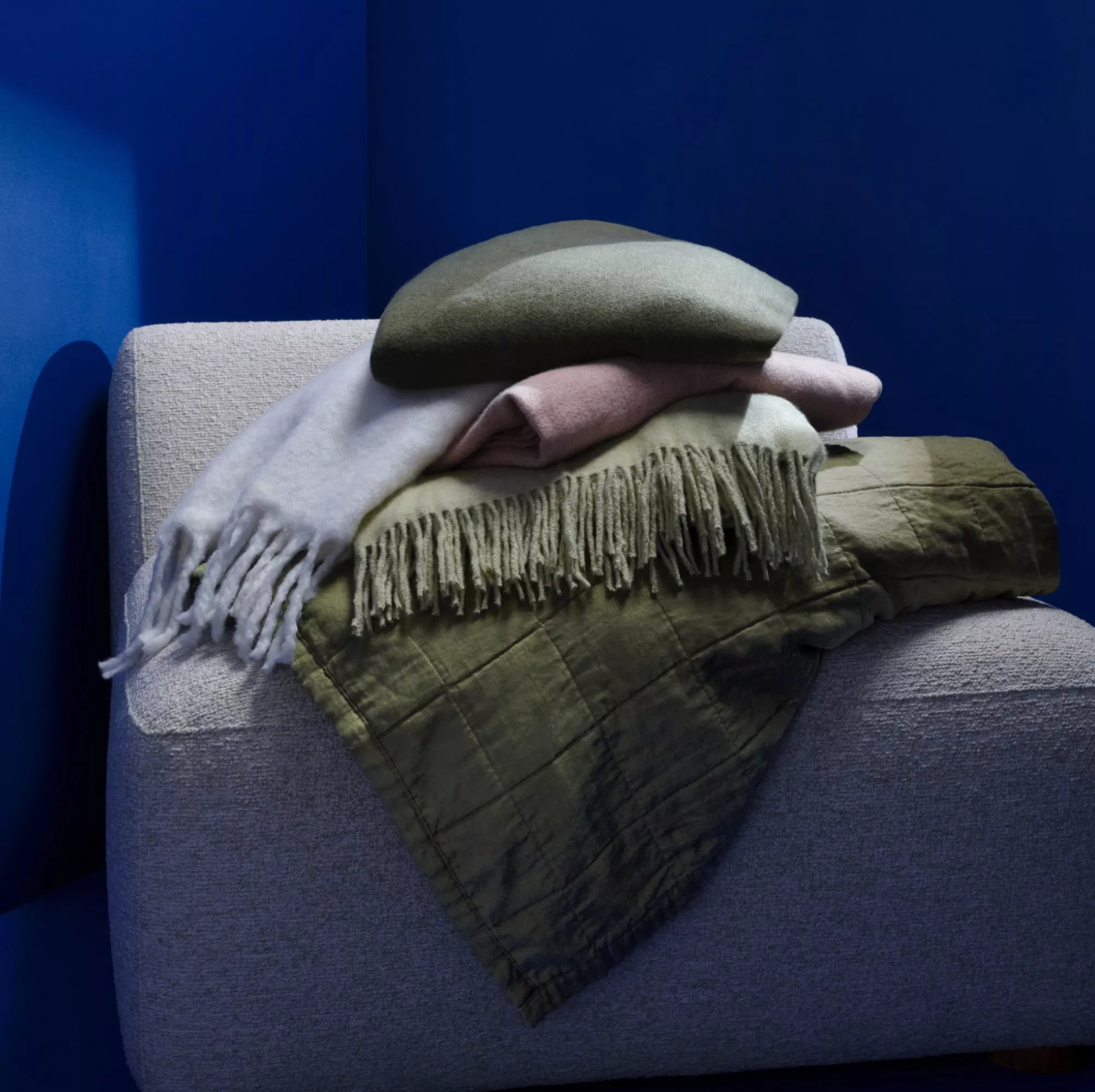 Stack of cosy throws and fringed blanket on grey armchair, blue wall backdrop, modern home décor styling