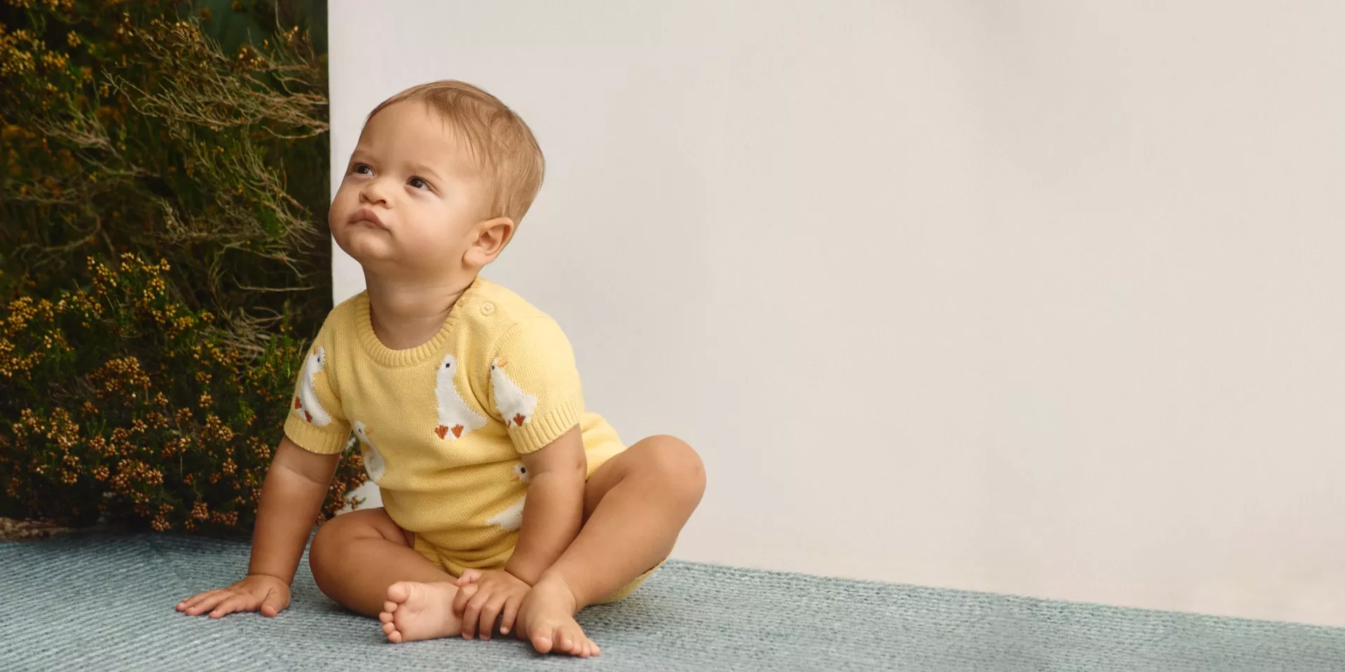 A toddler in a yellow outfit with white duck patterns sits on a blue rug next to greenery, looking upwards.