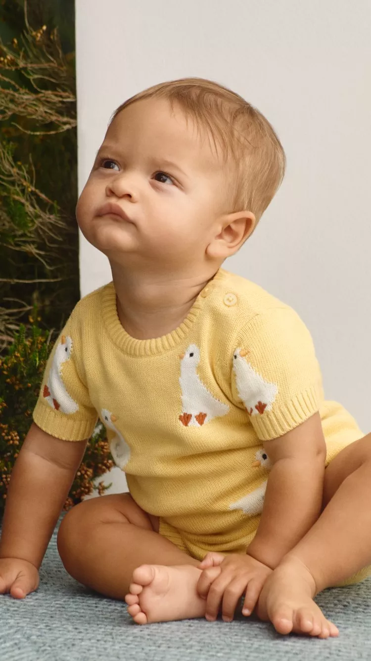 A baby wearing a yellow knit outfit with duck patterns sits on a blue carpet, surrounded by green foliage.