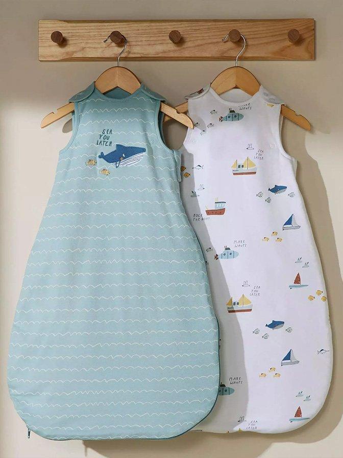 The image shows two sleeveless baby sleeping bags hanging on wooden hangers. One is teal with a whale and 