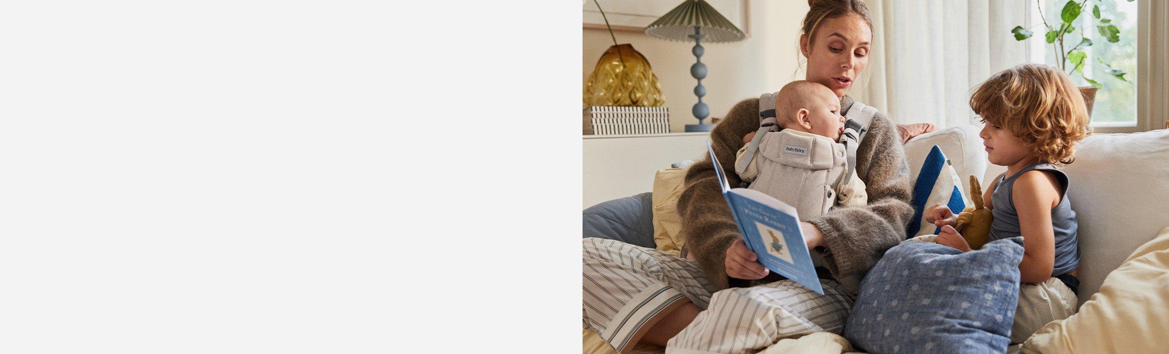 A woman with a baby in a front carrier and a young child sitting on a sofa, reading a book together in a cozy living room