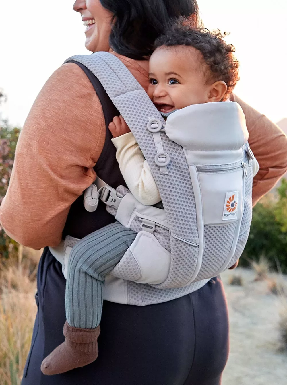 A woman carries a smiling baby in a grey baby carrier during an outdoor walk. The baby looks content and happy.