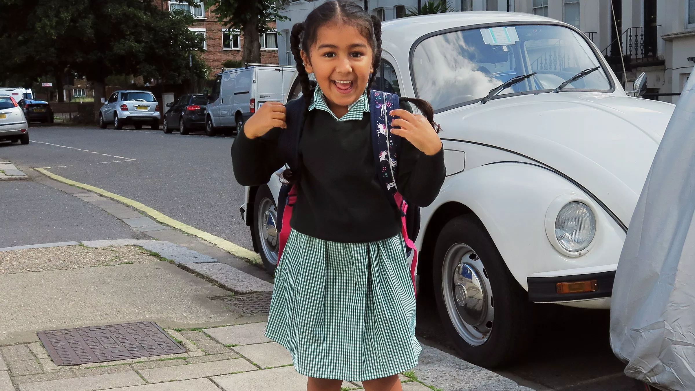 Smiling schoolgirl with backpack on a residential street, standing in front of a white classic Volkswagen Beetle car.