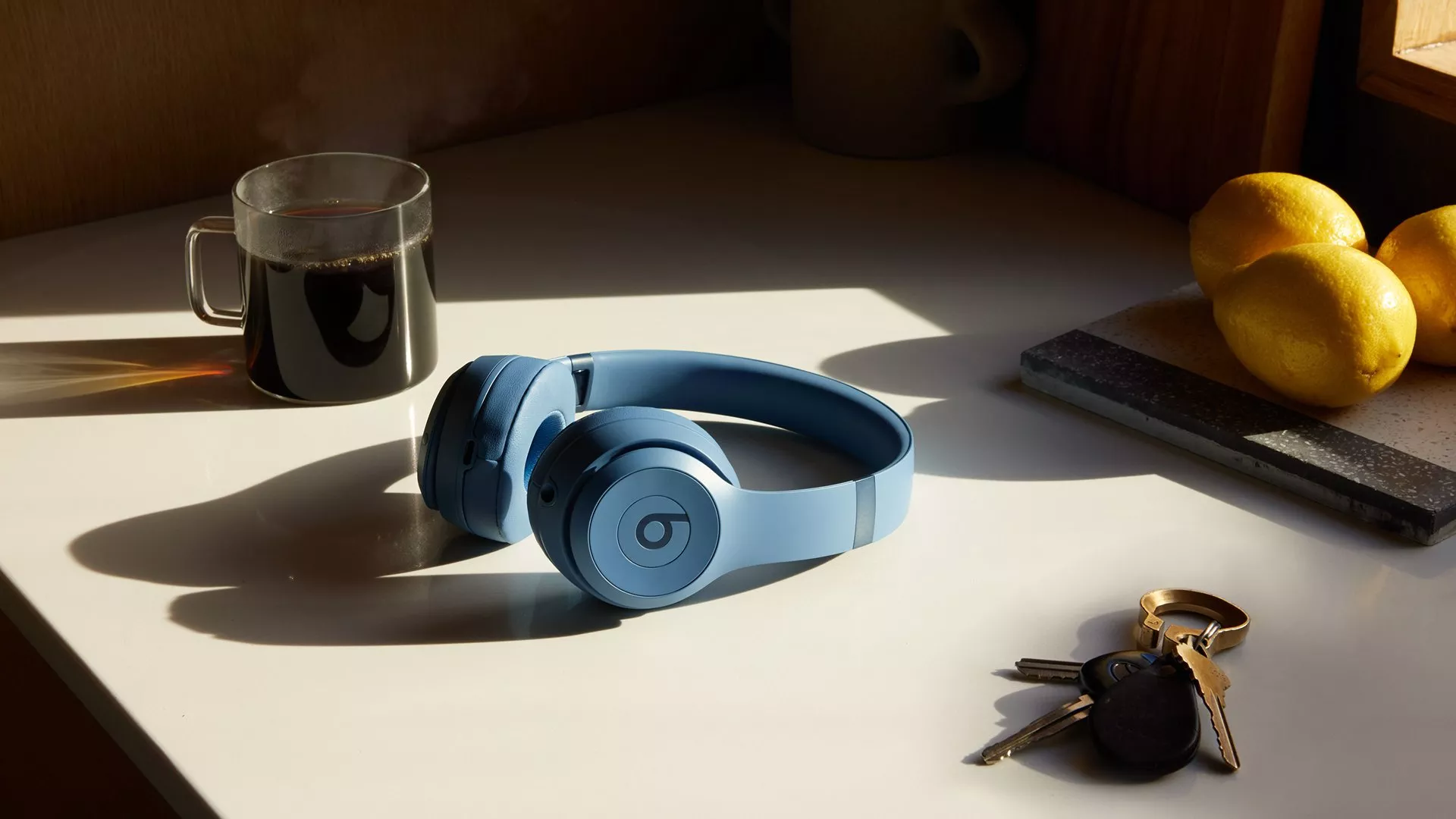 Blue over-ear wireless Beats headphones on kitchen counter with coffee mug, lemons and keys in warm sunlight