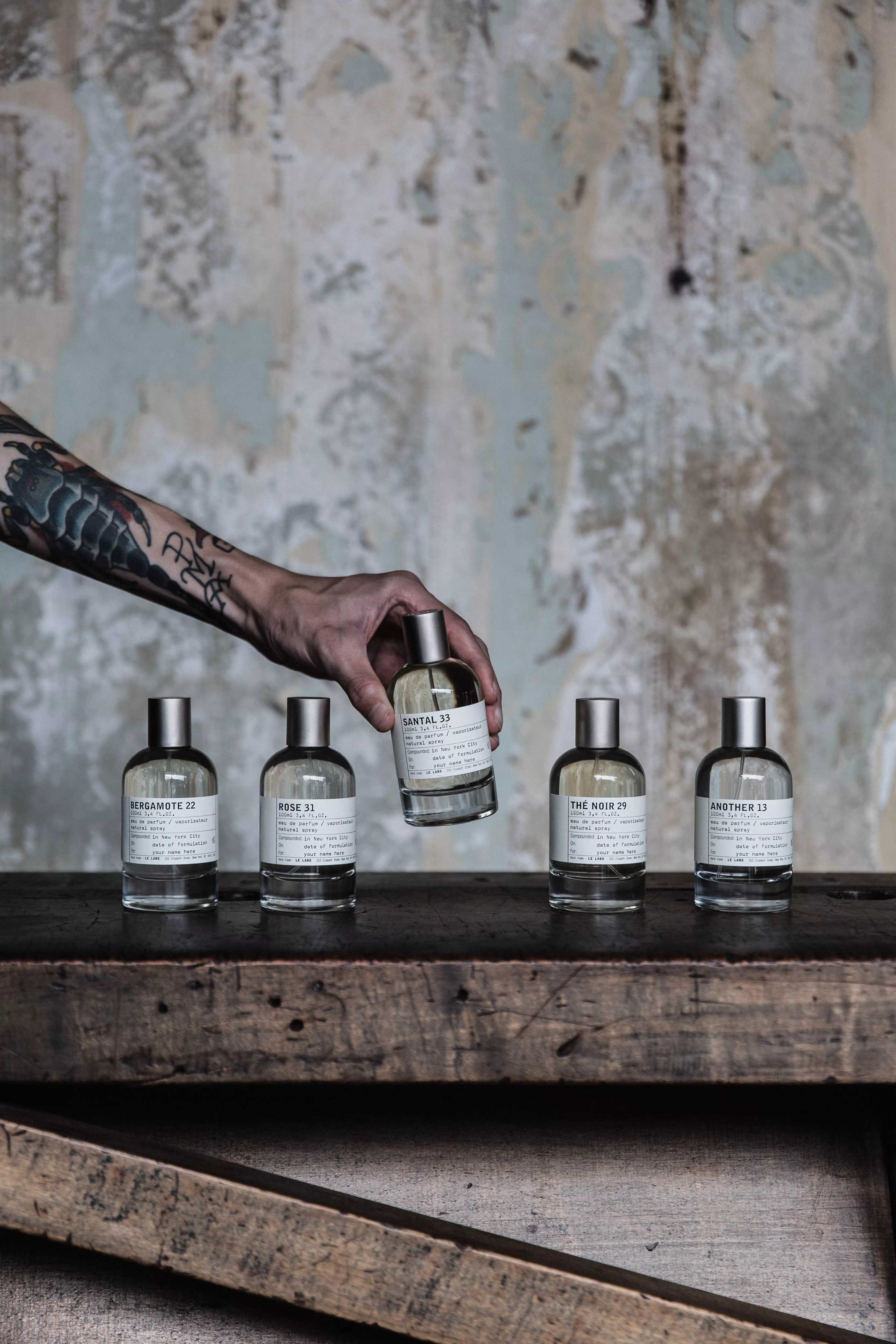  A tattooed arm reaches down to select a bottle of Santal 33 from a line of five Le Labo fragrances displayed on a weathered wooden surface.