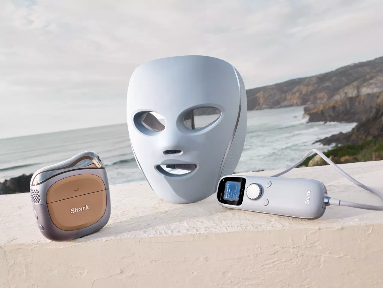 Shark LED face mask and skincare device set on a ledge, with ocean waves and coastal cliffs in the background