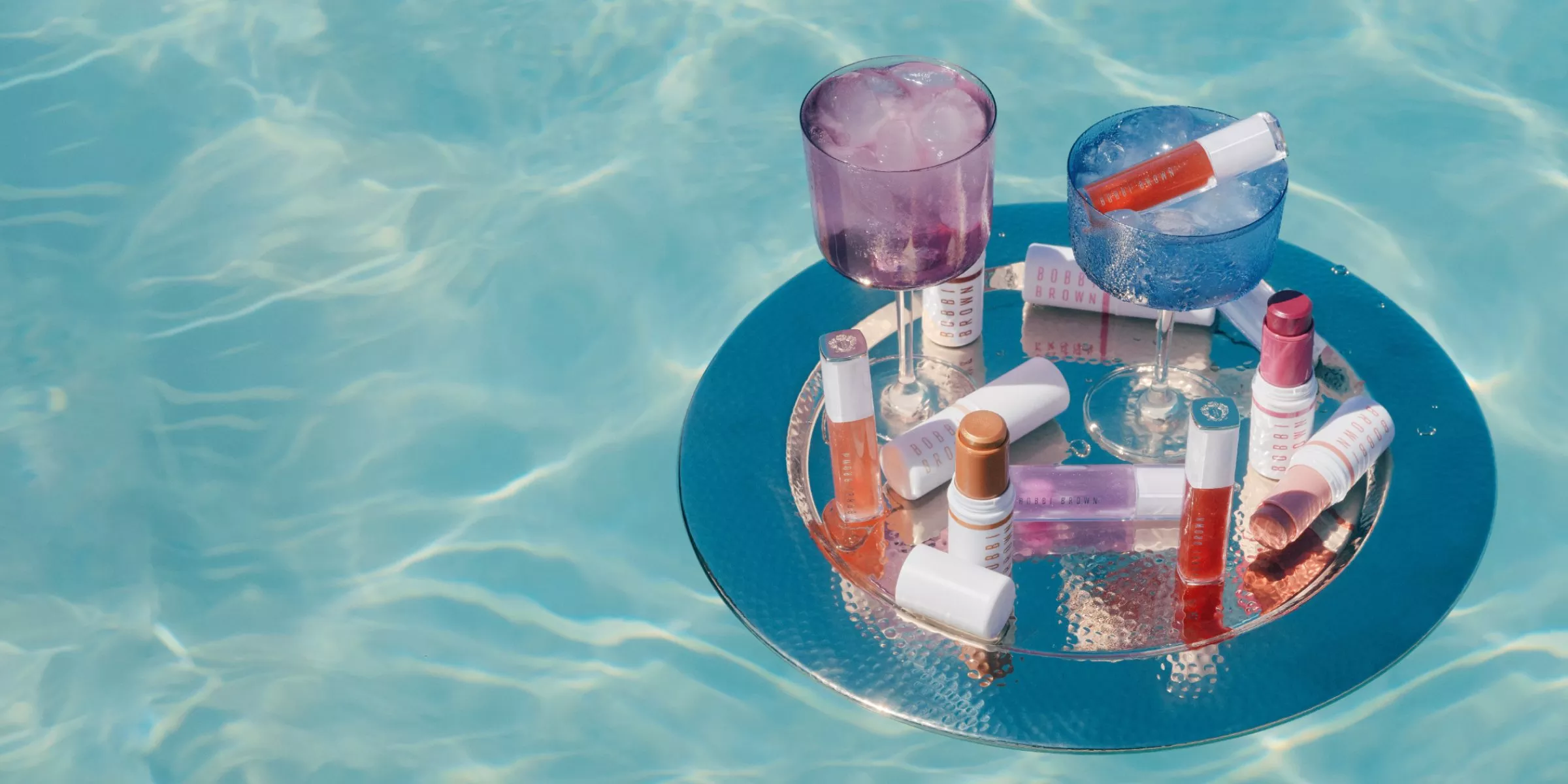 Luxury poolside beauty products and lip gloss on floating tray with colourful cocktail glasses in blue water