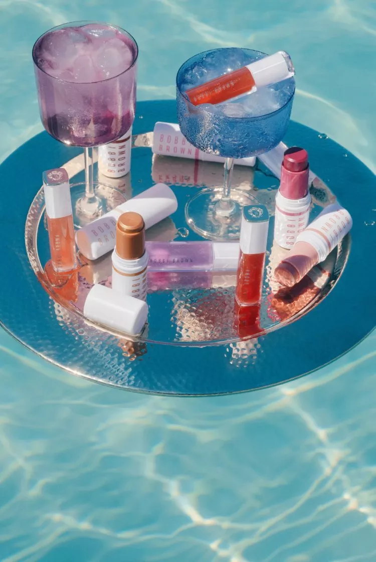 Summer poolside beauty flat lay with lip gloss, lipstick and makeup tubes on a silver tray with iced cocktails