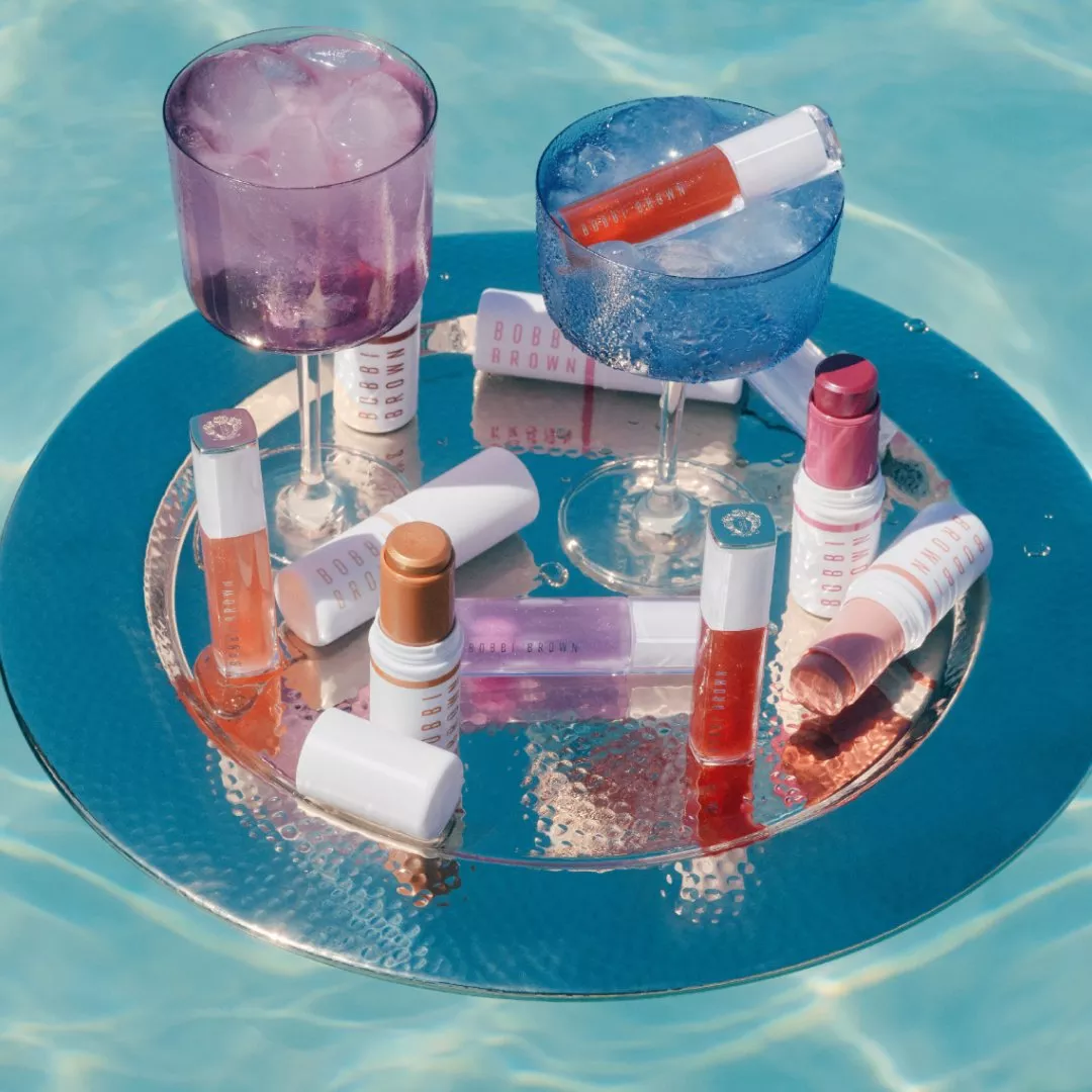 Bobbi Brown makeup and lip gloss on a silver tray floating in a swimming pool with iced cocktails