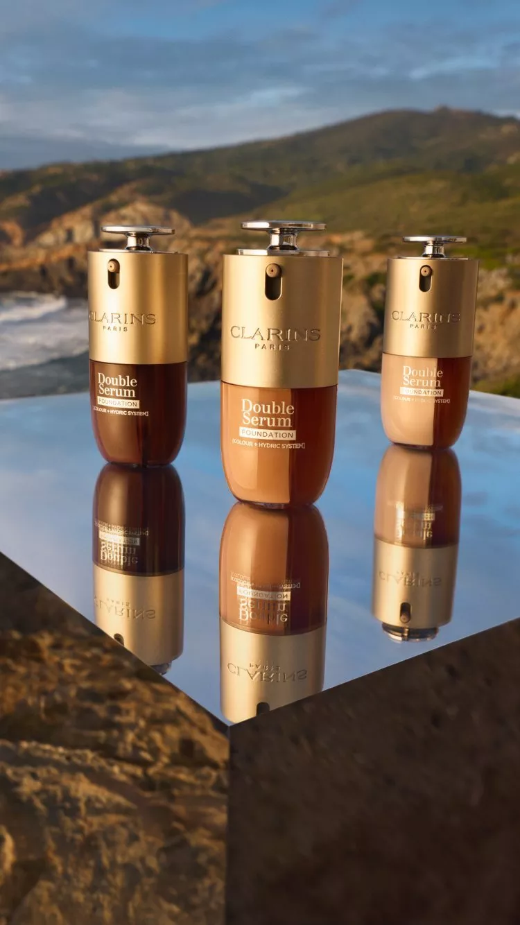 Clarins Double Serum bottles displayed on a mirrored surface outdoors, with scenic hills in the background.