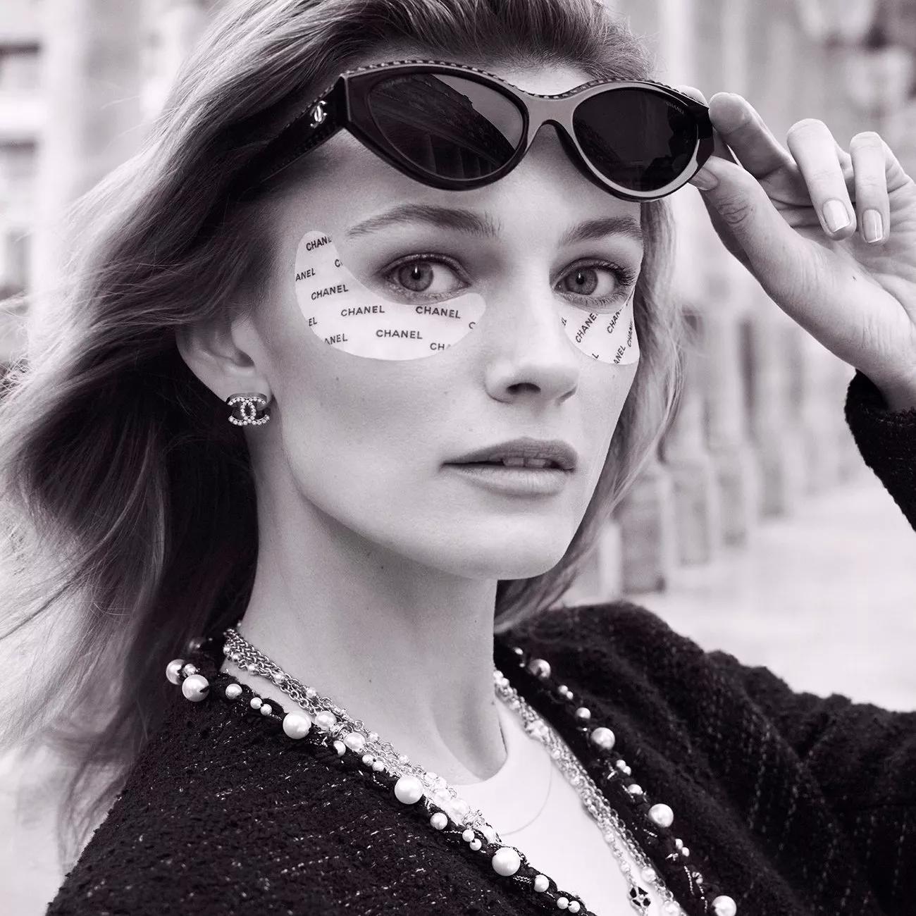 Stylish woman in monochrome wearing Chanel eye patches, sunglasses, and statement jewellery outdoors.