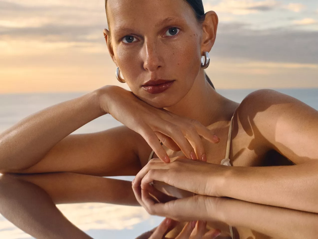 A serene sunset backdrop reflects a woman's hands resting on a surface, showcasing elegant jewellery.