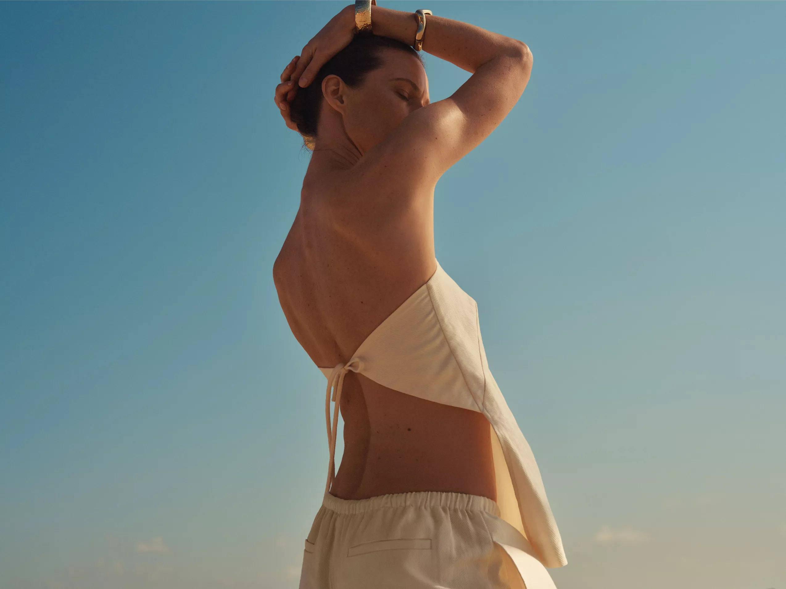 Back view of woman in cream summer outfit with arms raised, sunlit against clear blue sky outdoors.