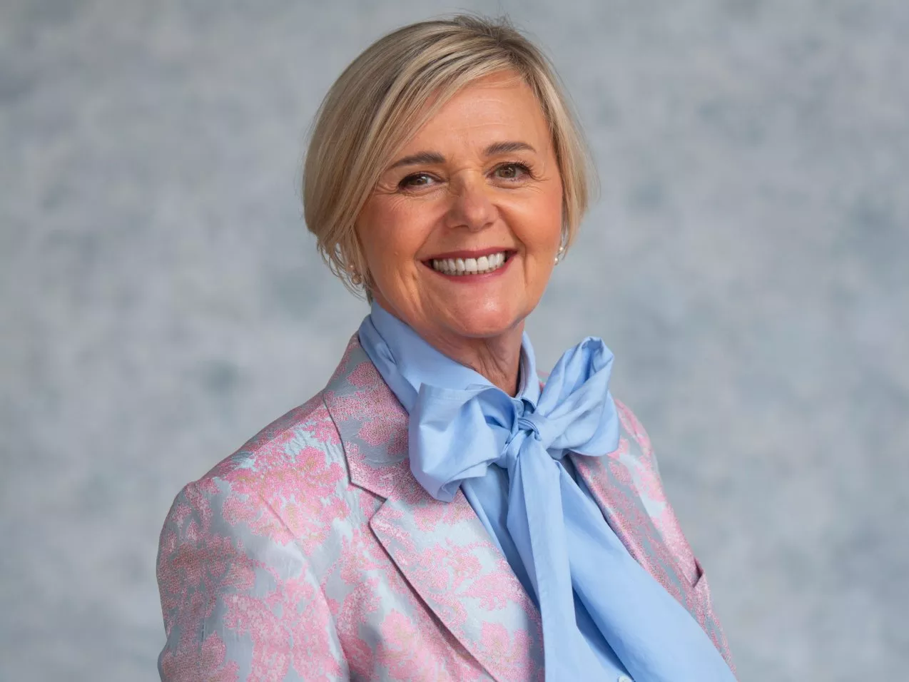 Smiling woman in a pink floral blazer with a large blue bow tie against a soft blue background.