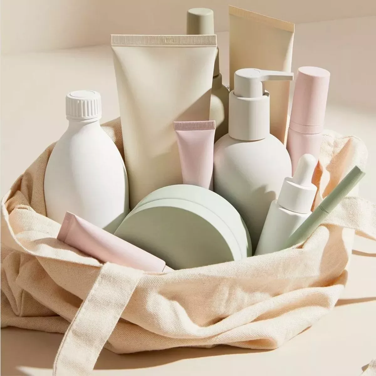 collection of unlabeled cosmetic and skincare products in various sizes and muted pastel colors (pale green, beige, and soft pink) contained within a reusable canvas tote bag.