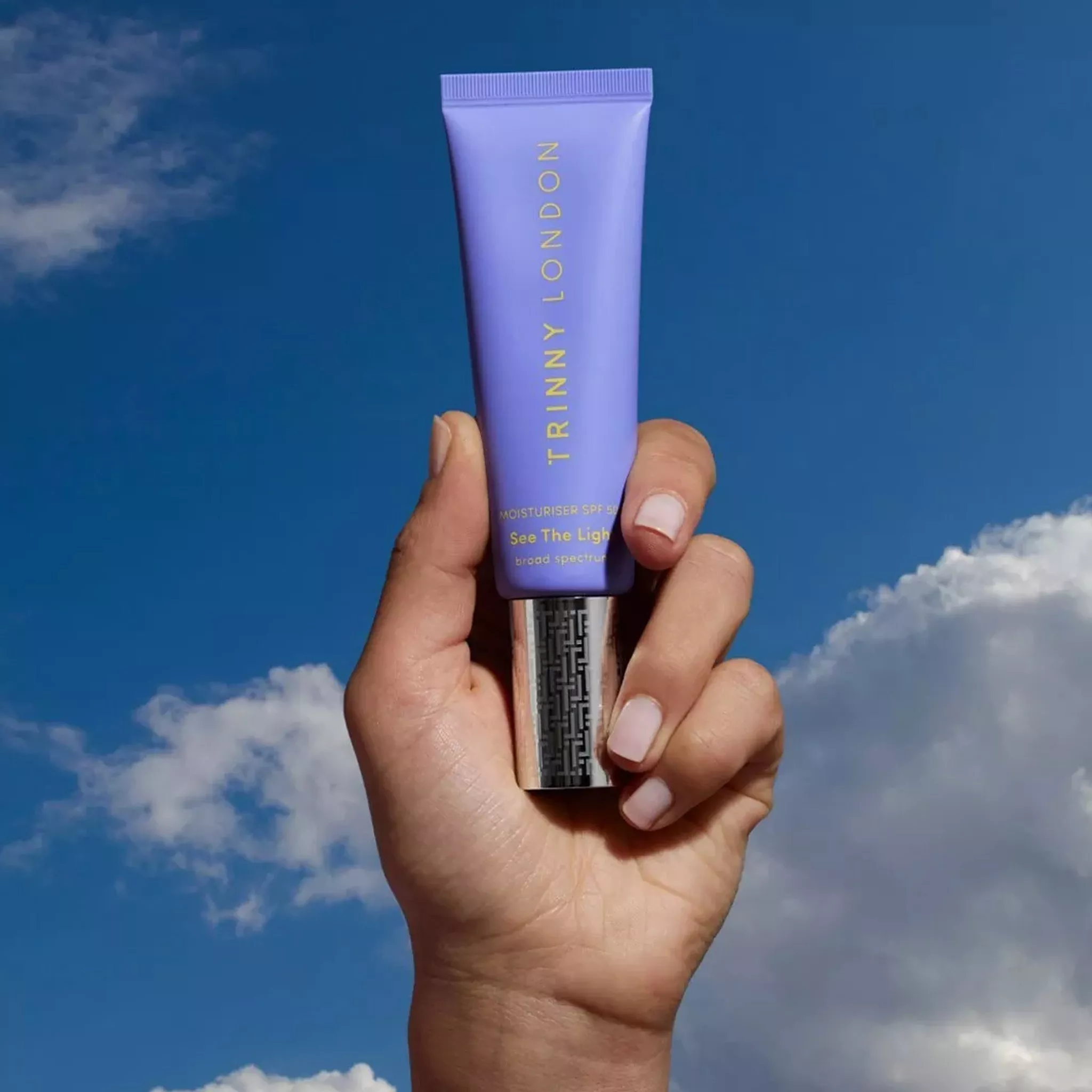 Hand holding Trinny London See The Light moisturiser SPF 50 tube against blue sky with clouds background