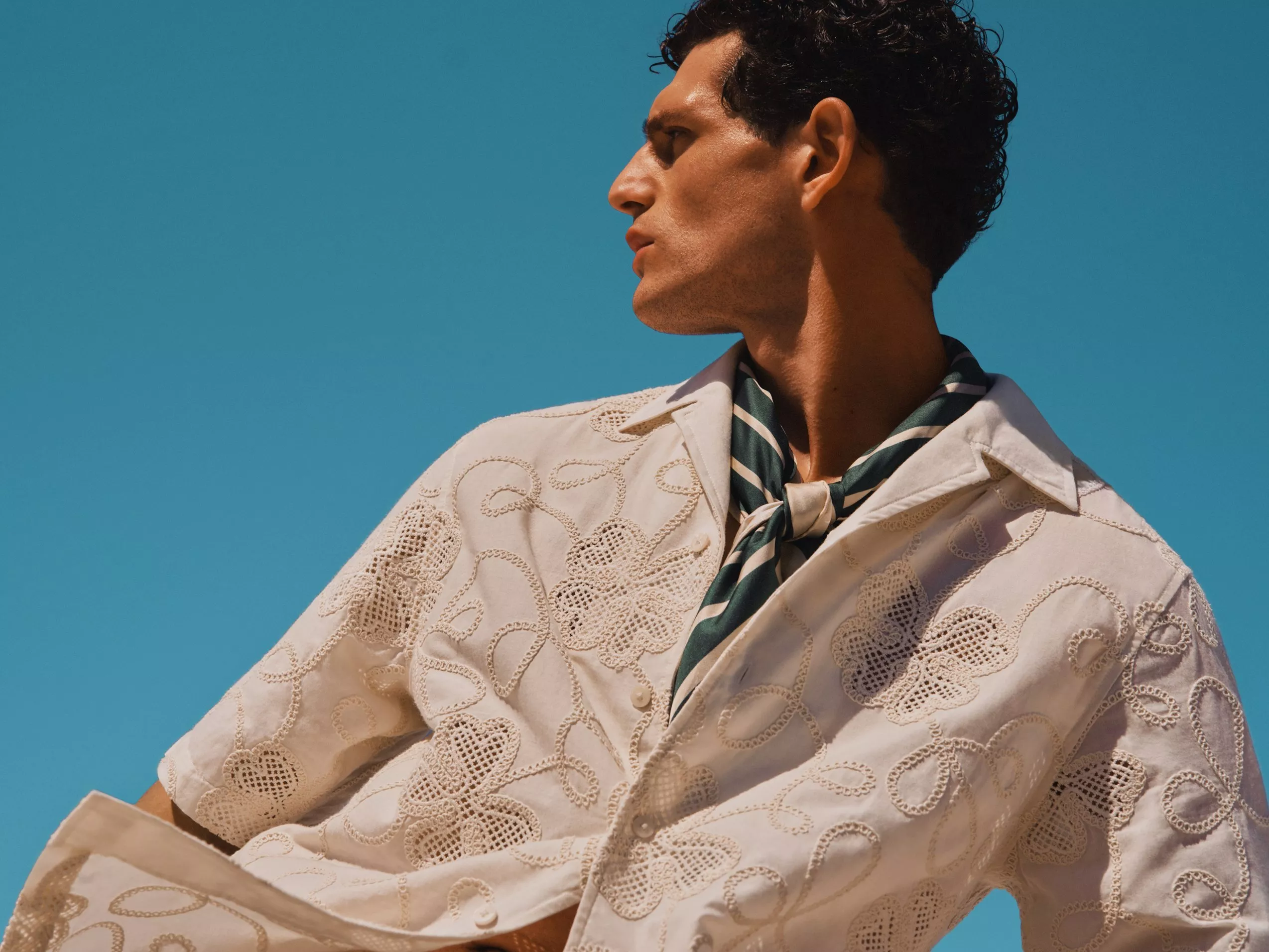 Side profile of man in embroidered white shirt and striped neck scarf against clear blue sky, summer fashion portrait