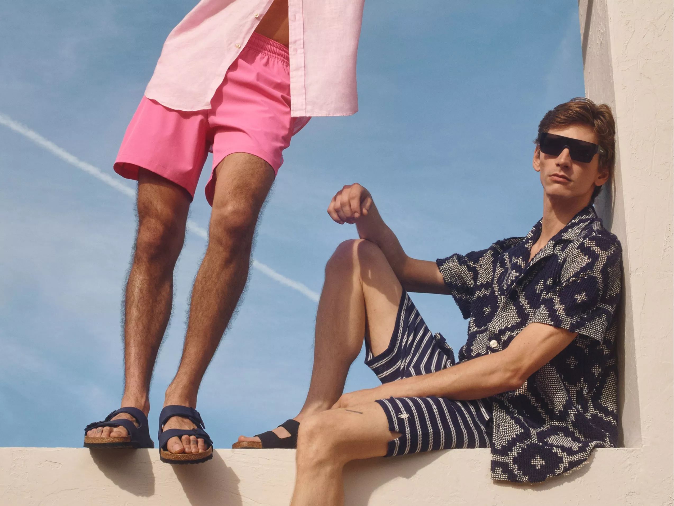 Two men modelling summer outfits with shorts, sandals and sunglasses against a bright blue sky backdrop
