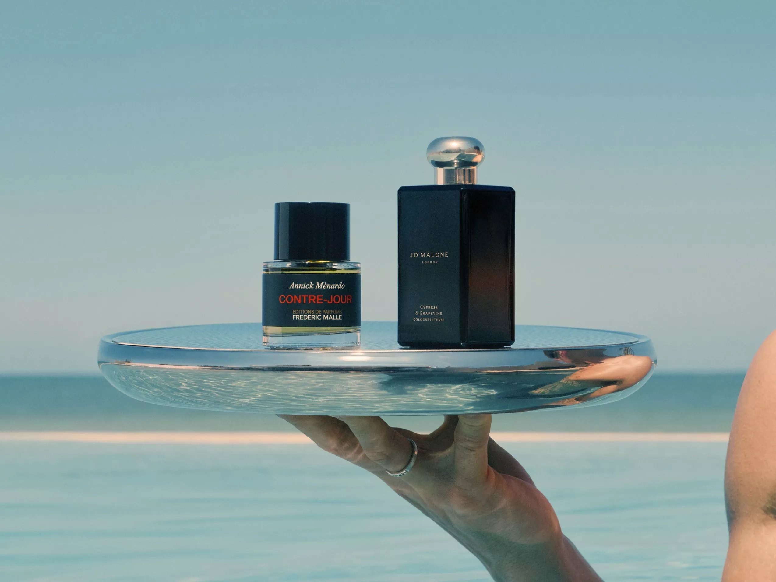 Jo Malone and Frederic Malle perfumes bottles atop a hand held silver tray by the sea