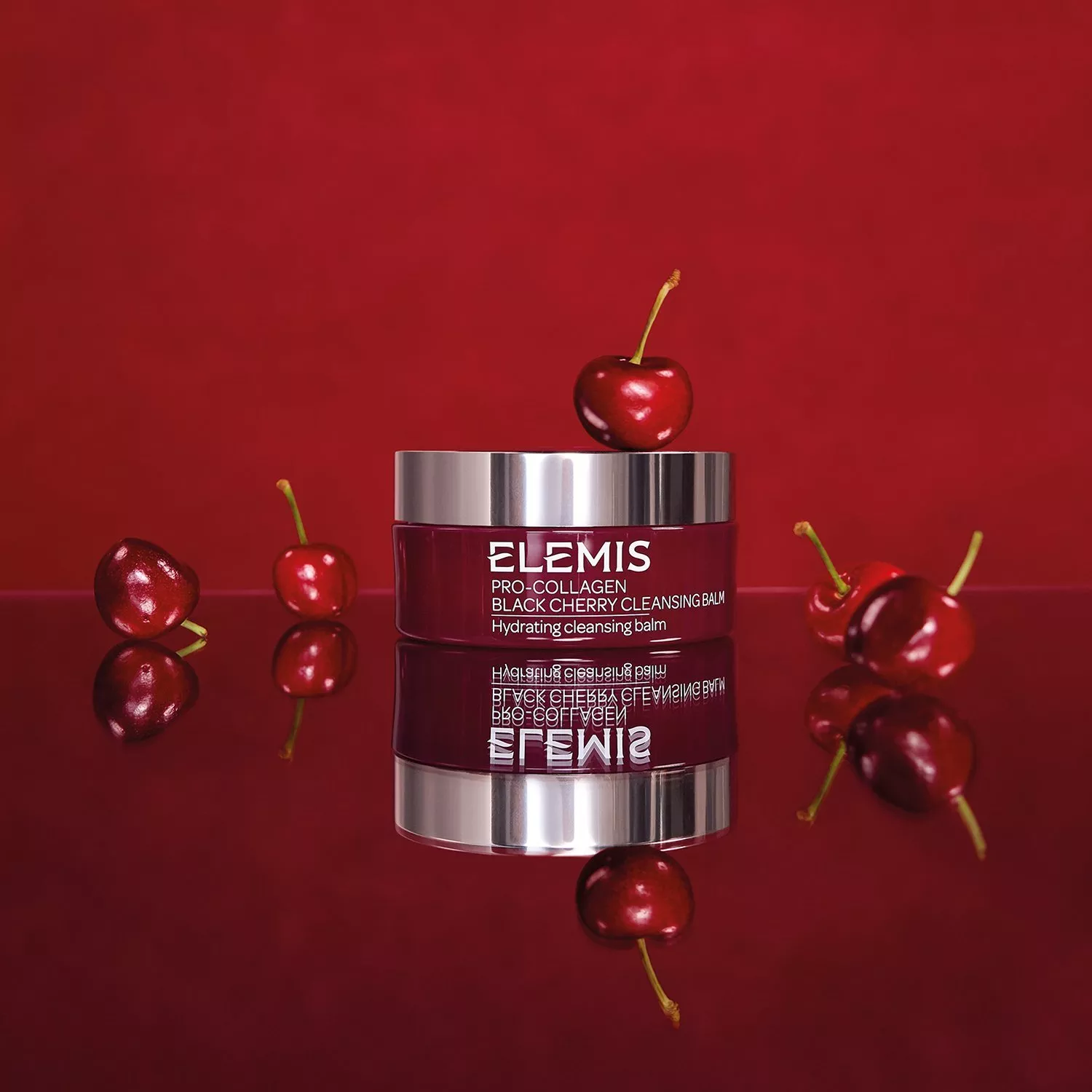 Image of an Elemis Pro-Collagen Balm on a red reflective backdrop with cherries around it