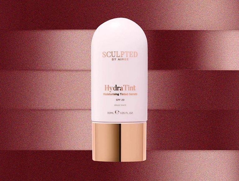 Assorted luxury beauty products including a Charlotte Tilbury blush, Clinique serum, Estée Lauder lipstick, and Chantecaille cream on a pink background.