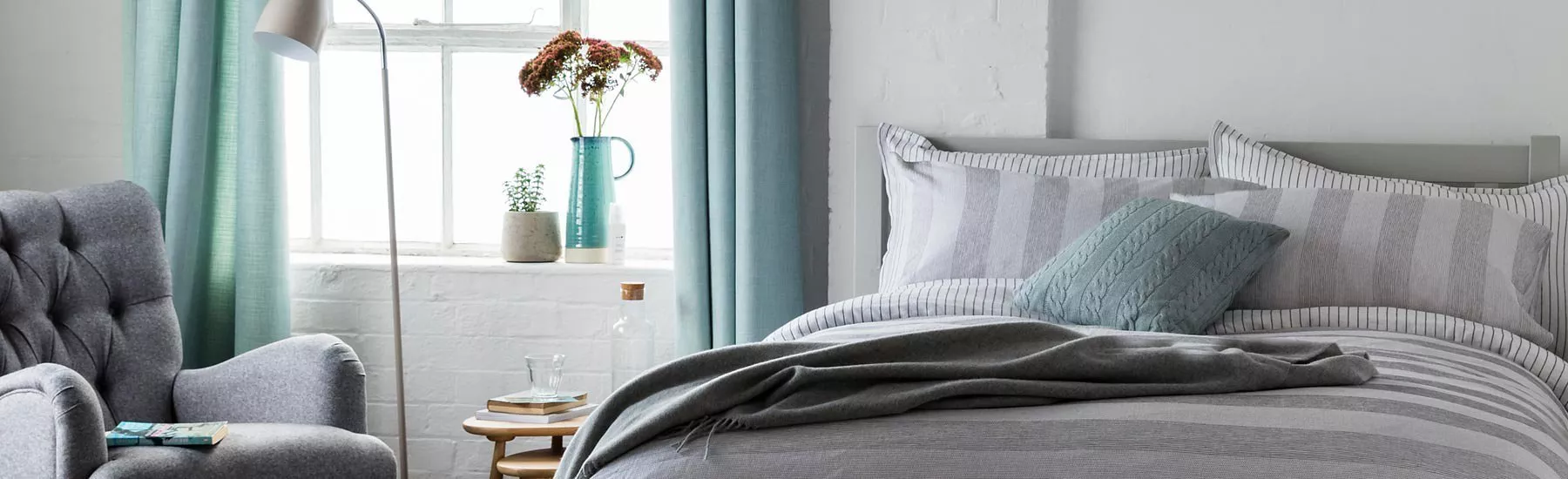 Modern bedroom with grey striped bedding, teal cushions and curtains, armchair, floor lamp and window light