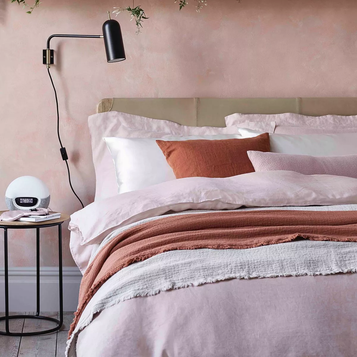 Modern bedroom with pink linen bedding, rust throw, wall-mounted black reading lamp and bedside table decor