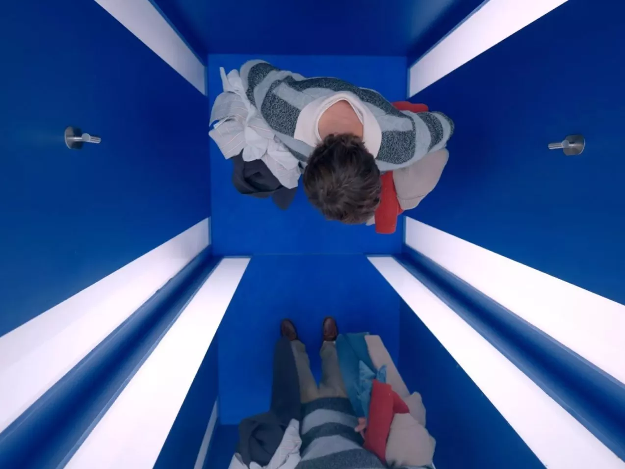 Overhead view of two people in a blue changing room cubicle holding clothes, bright white strip lighting.