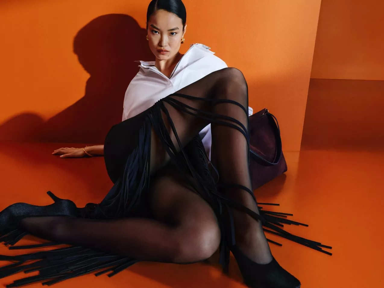 Fashion model in white shirt and black sheer tights posing on orange studio backdrop with handbag