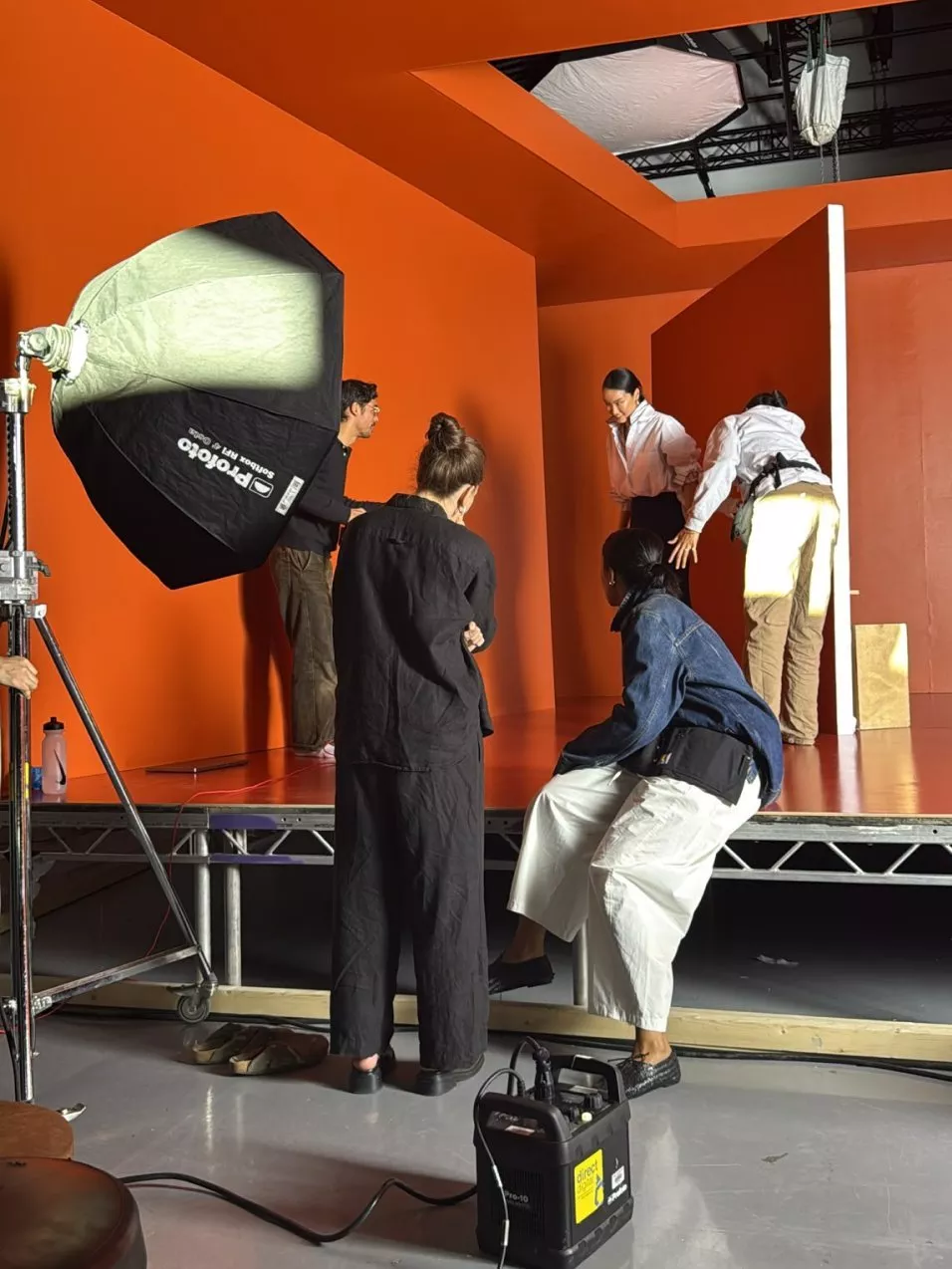 Fashion photoshoot set with crew and model on orange backdrop, studio lighting and softbox behind the scenes