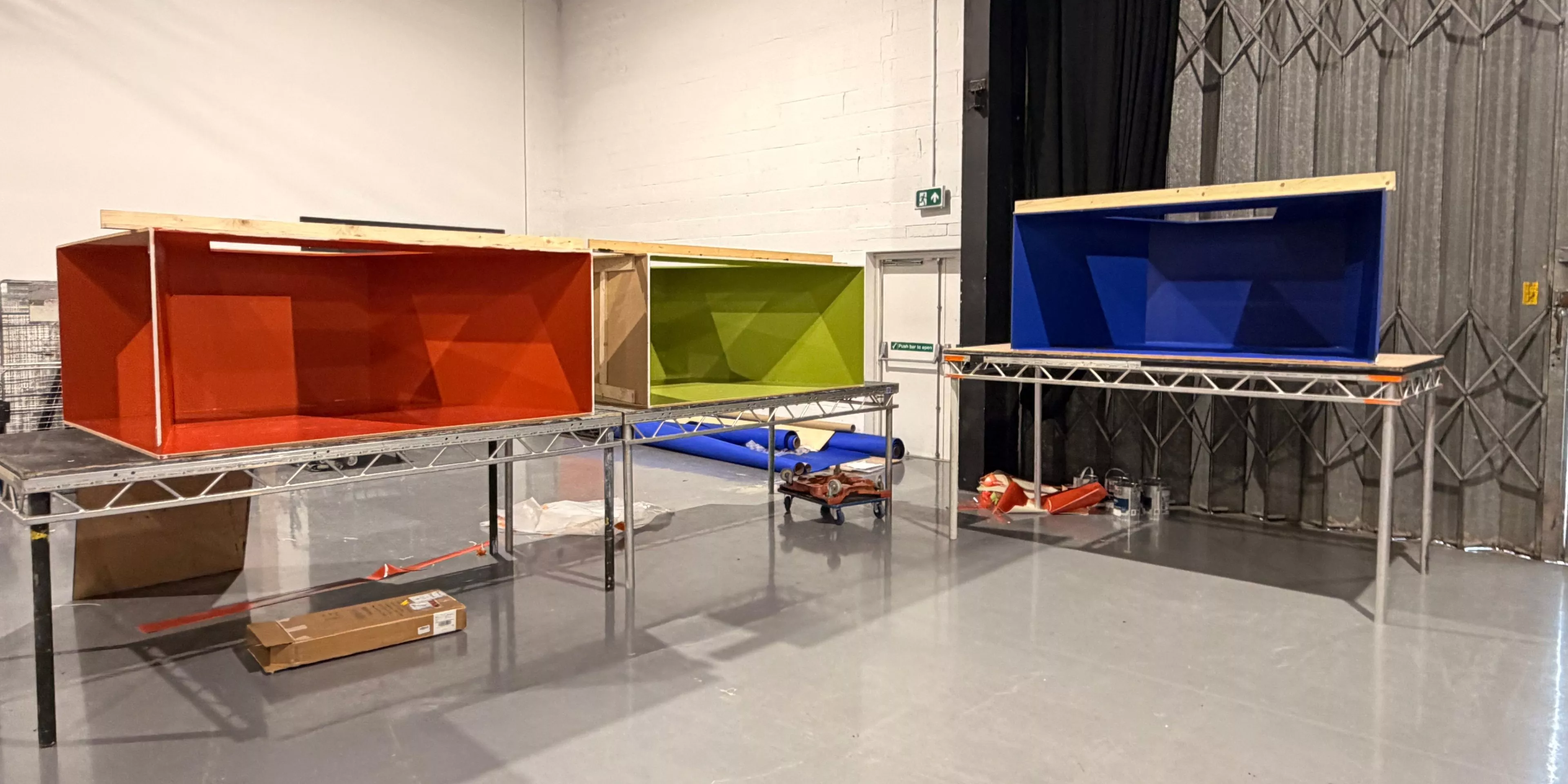 Three large colourful wooden display boxes on metal tables in a warehouse workshop, red, green and blue units.