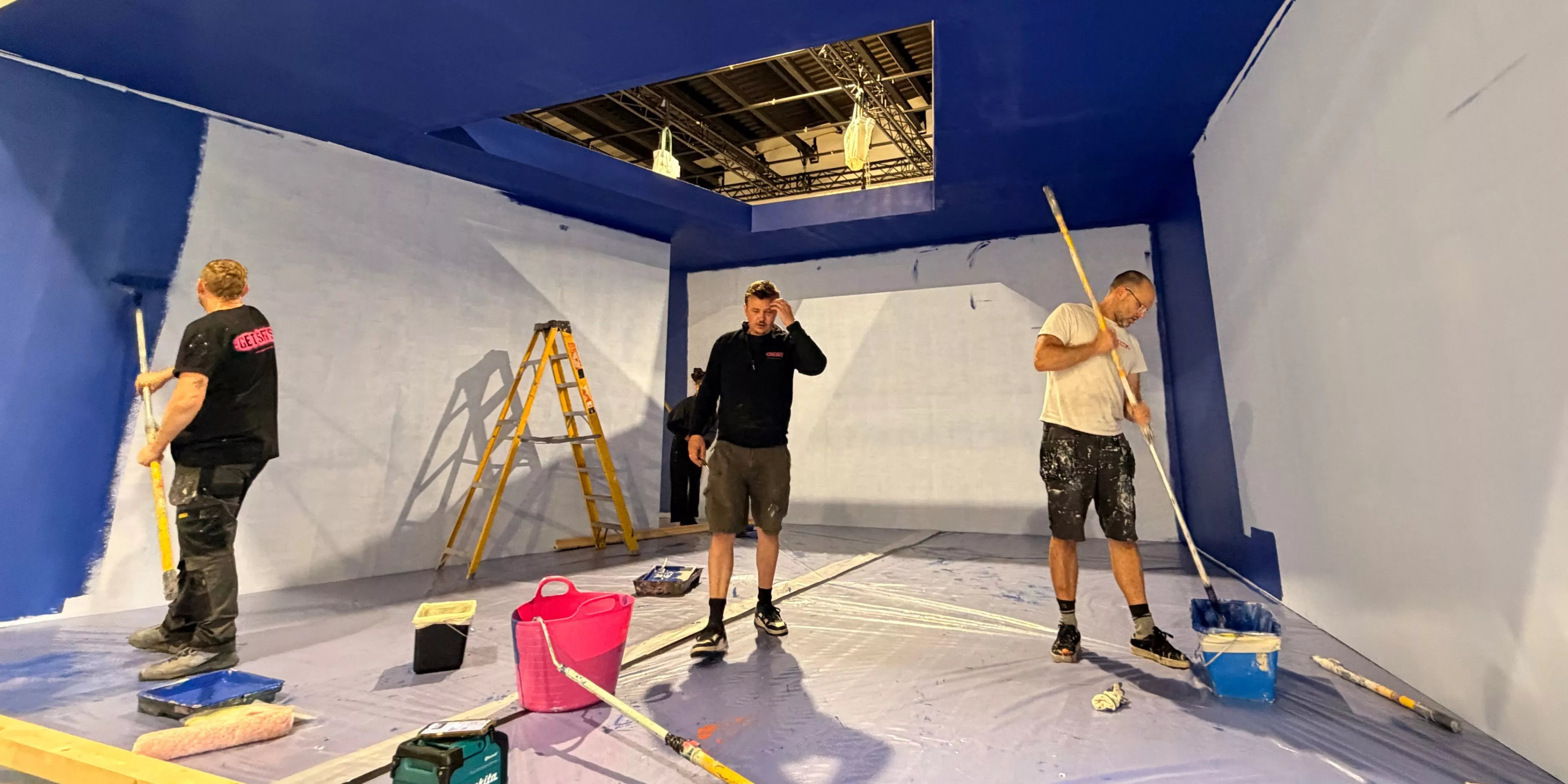 Commercial painters roller-applying blue paint to interior walls and ceiling, with ladder and tools on site