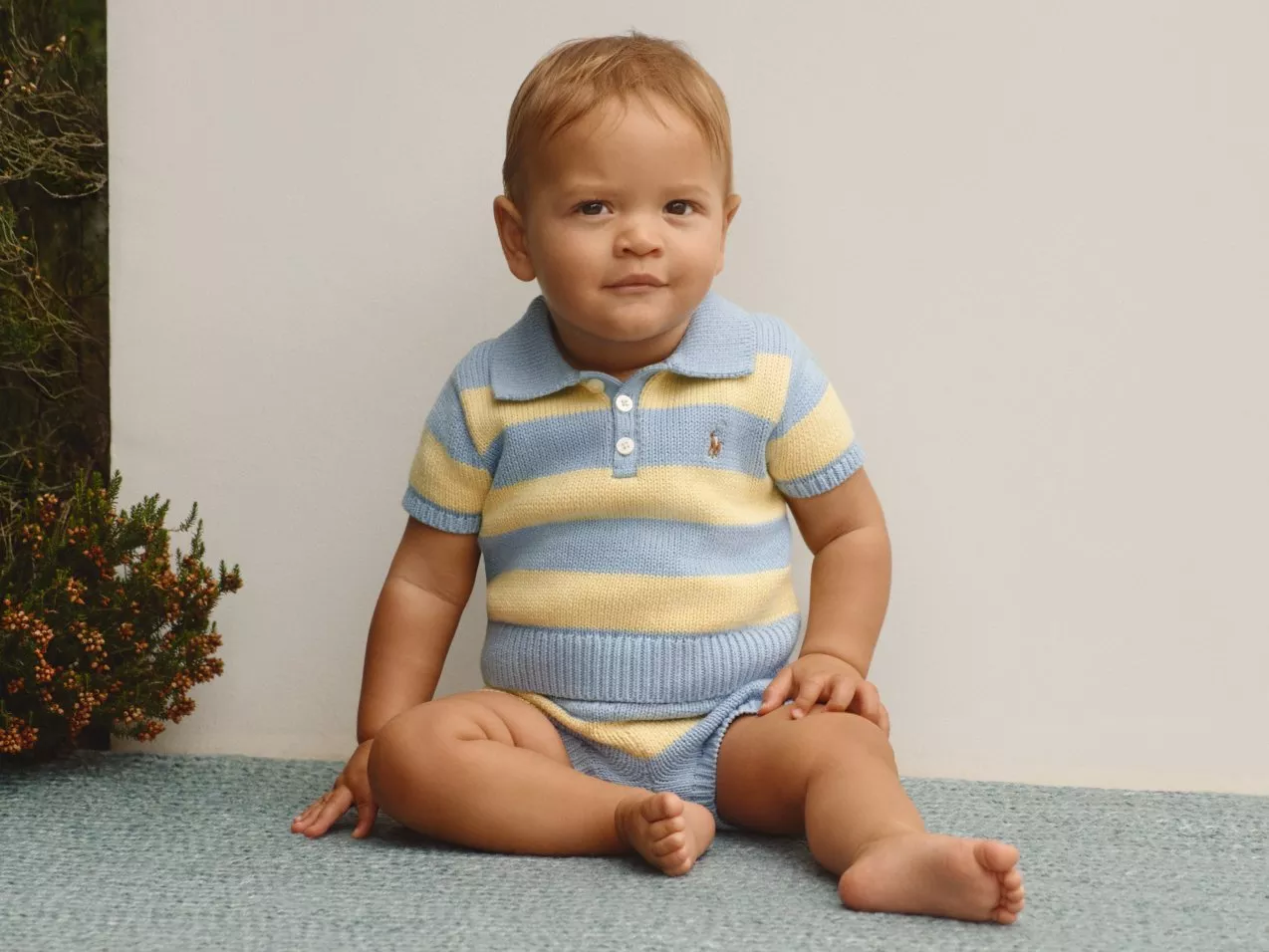 Baby boy sitting on carpet in blue and yellow striped knit romper, studio portrait with neutral backdrop