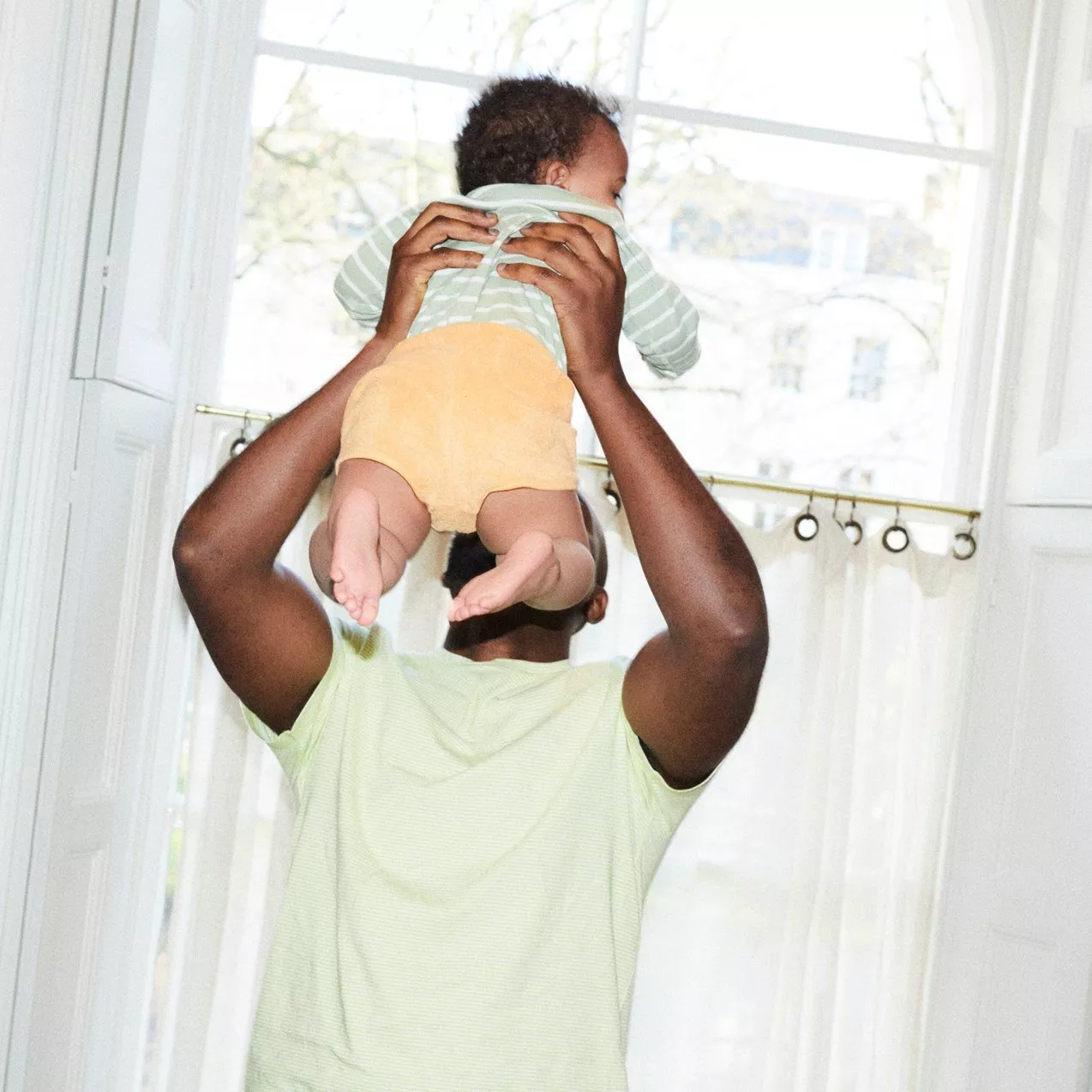 Father lifting baby in bright home by window, playful bonding moment and family lifestyle indoors