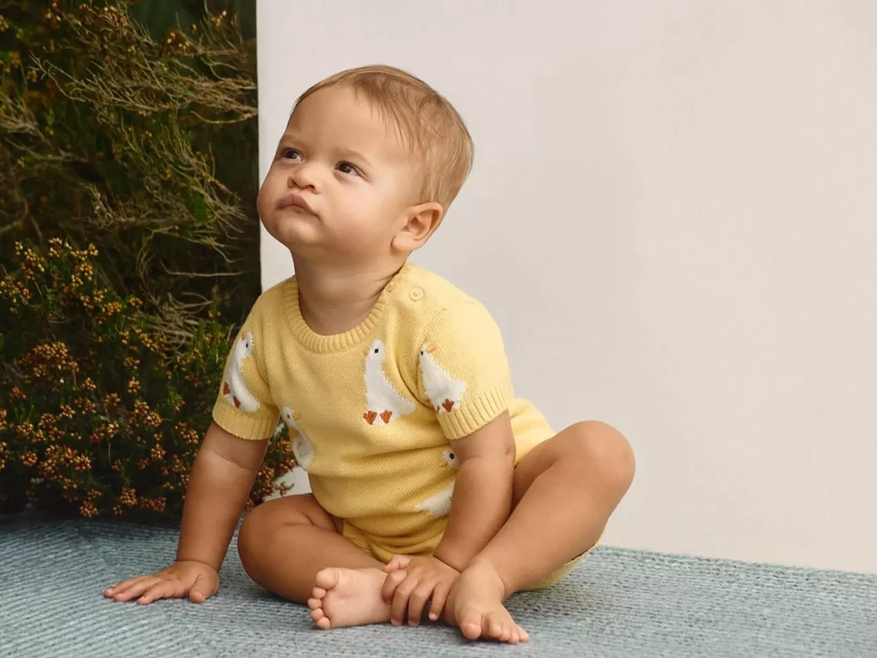 Baby sitting on blue rug wearing yellow knitted romper with duck pattern, looking up in studio setting