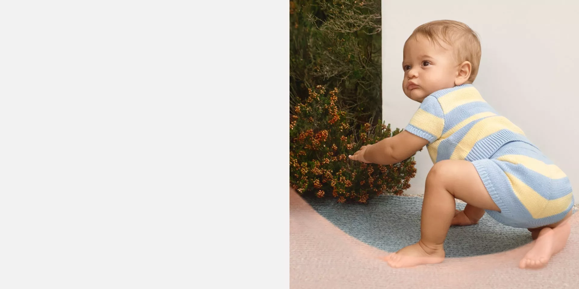 Baby crawling outdoors in a blue and yellow striped knitted romper beside green shrubs on a patio rug