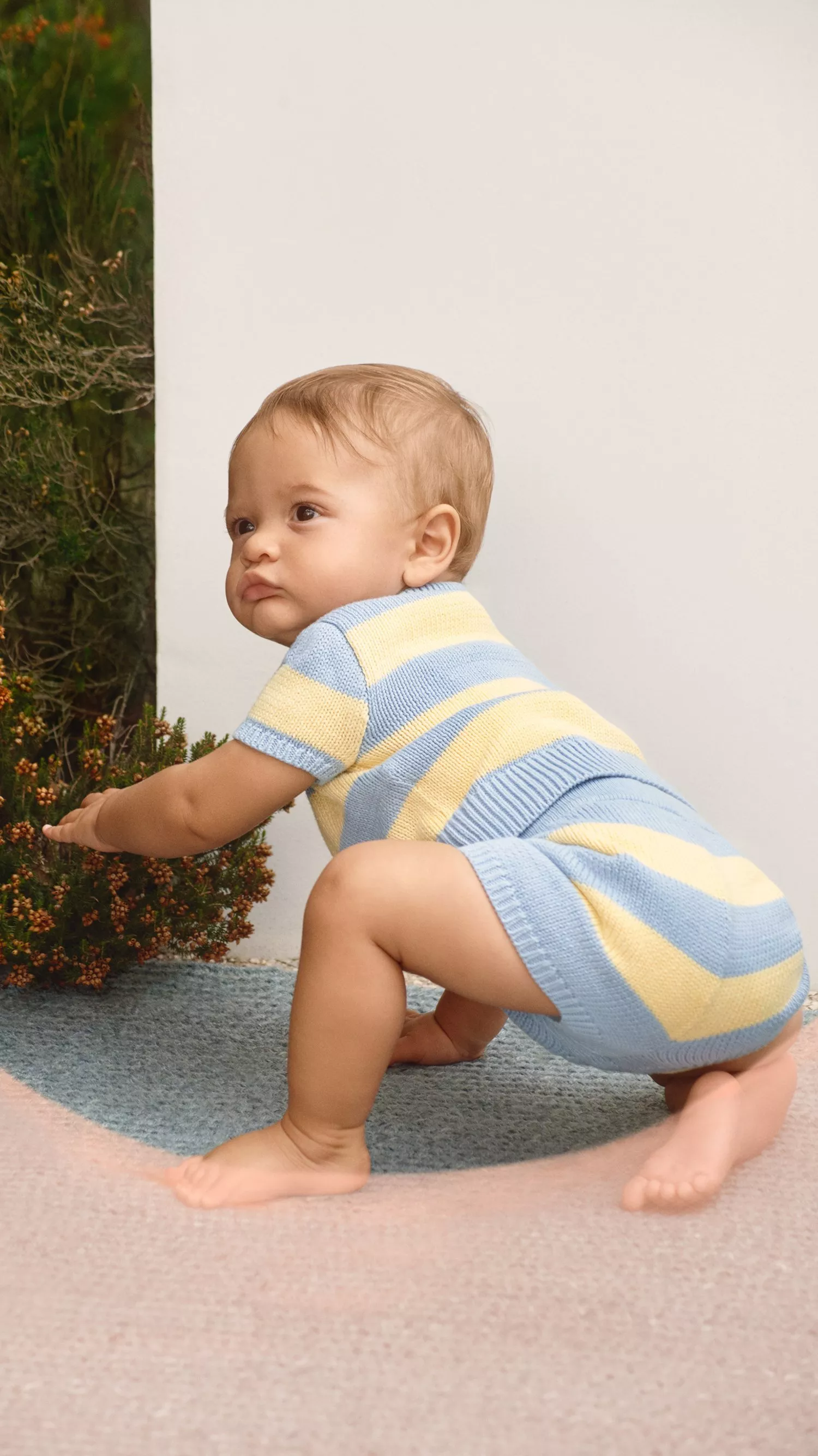 Baby crawling outdoors in a blue and yellow striped knitted romper beside green shrubs on a patio rug
