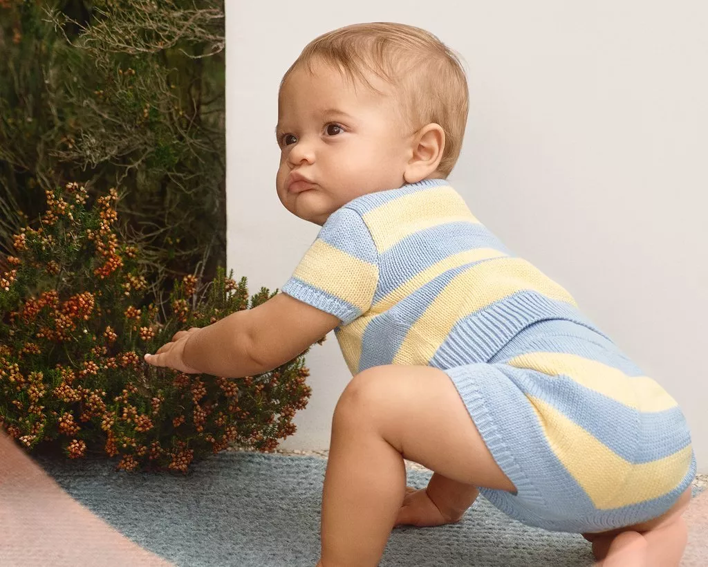Toddler in blue and yellow striped knitted romper crawling on a patio outdoors beside shrubs