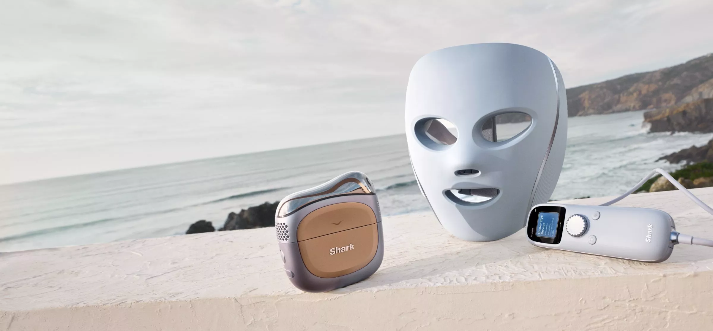 Three skincare devices on a ledge by the sea, including a futuristic mask and two electronic gadgets.