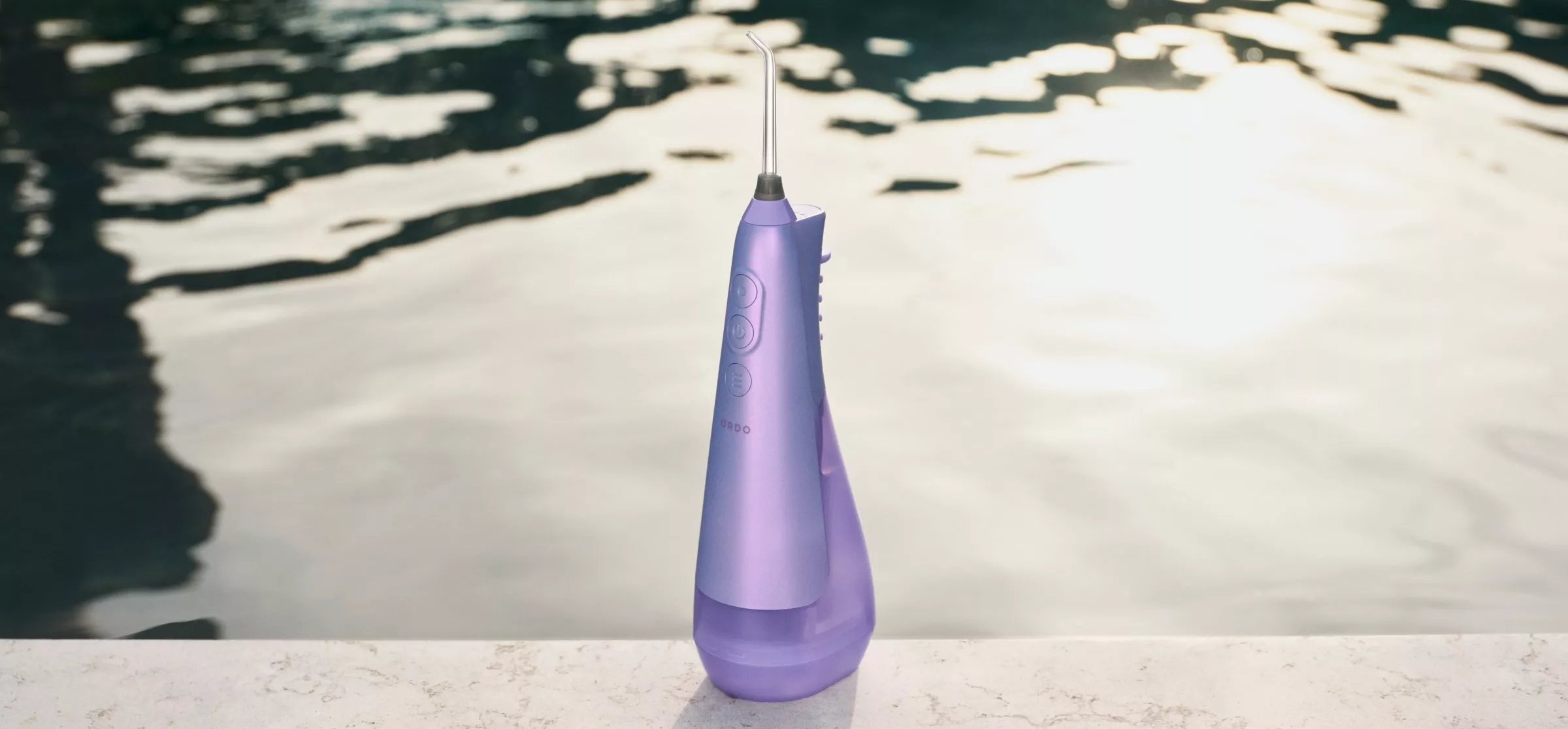 Purple water flosser with sleek design, placed on a stone surface by a serene pool, reflecting sunlight.